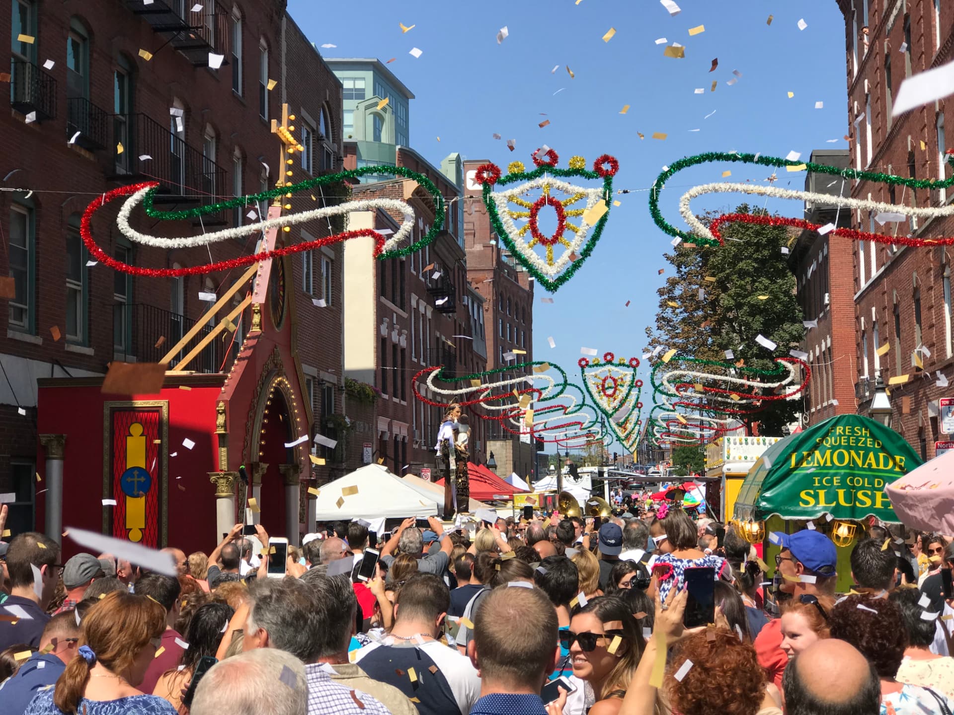 A crowded summer street festival with confetti, colorful garlands, and a procession float beneath a clear blue sky