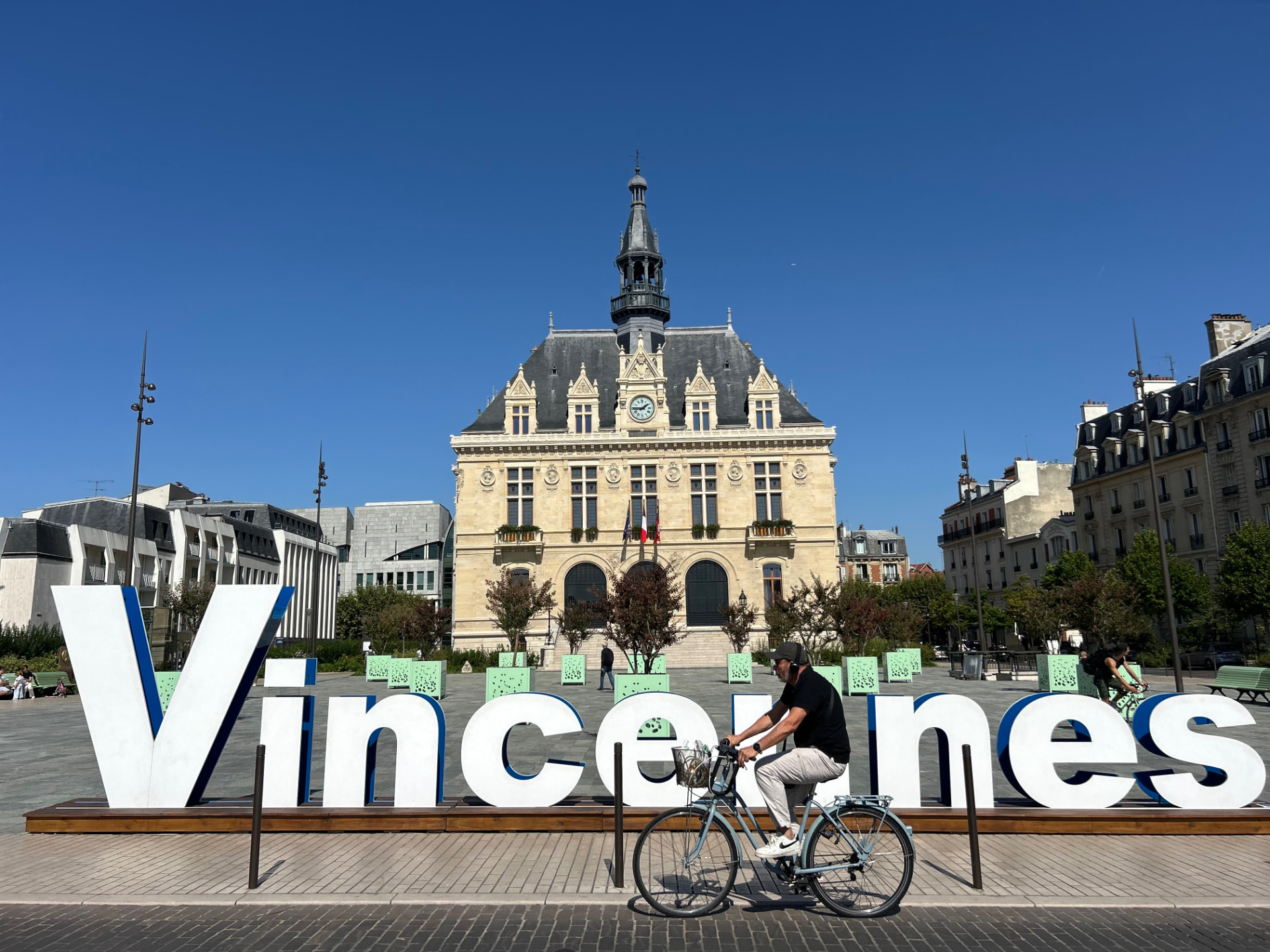 La mairie de Vincennes vue depuis la place, avec les grandes lettres Vincennes au premier plan et un cycliste qui passe sous un ciel bleu