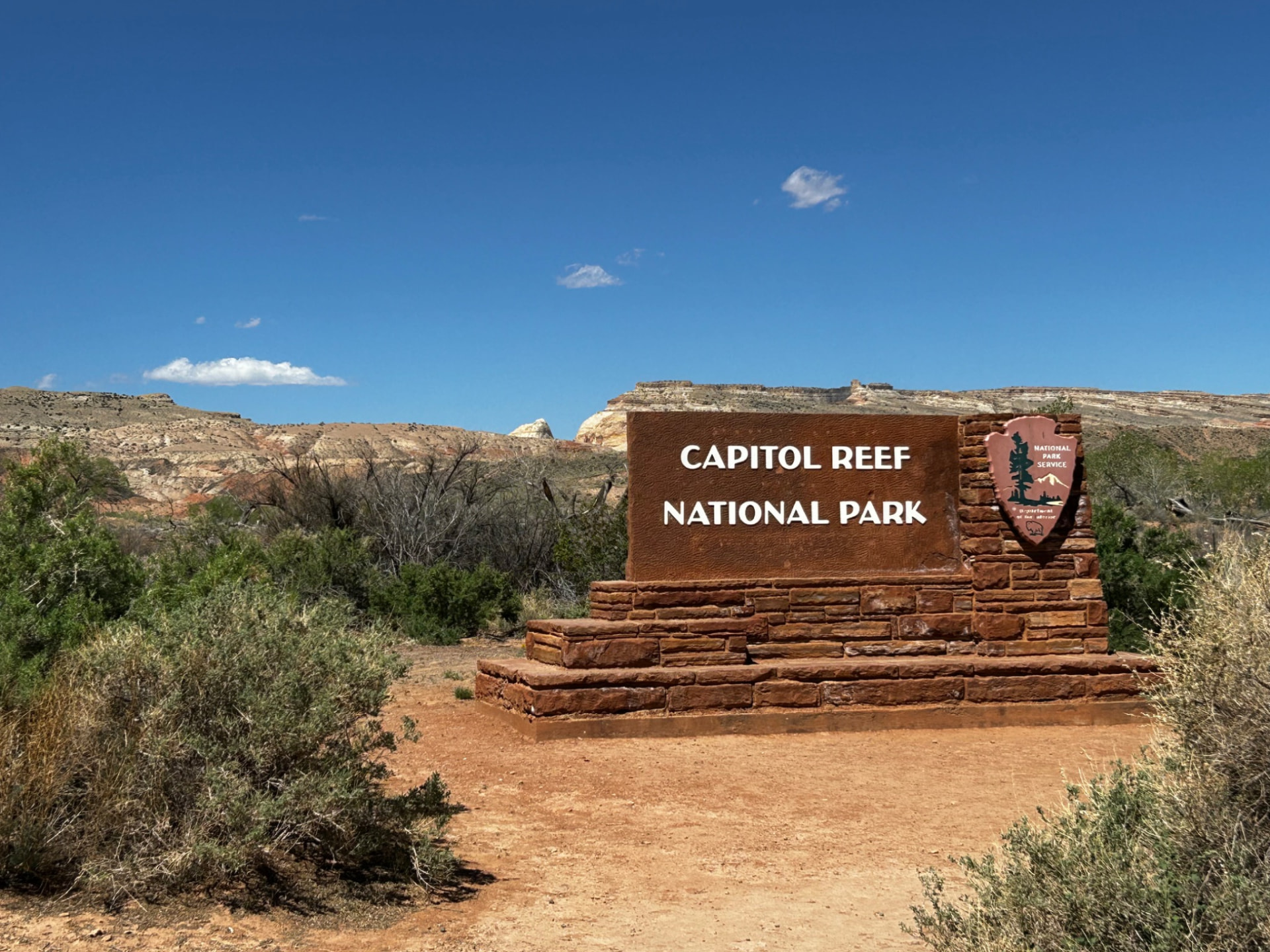 Capitol Reef National Park entrance sign in red stone, with desert shrubs, cliffs and blue sky behind it